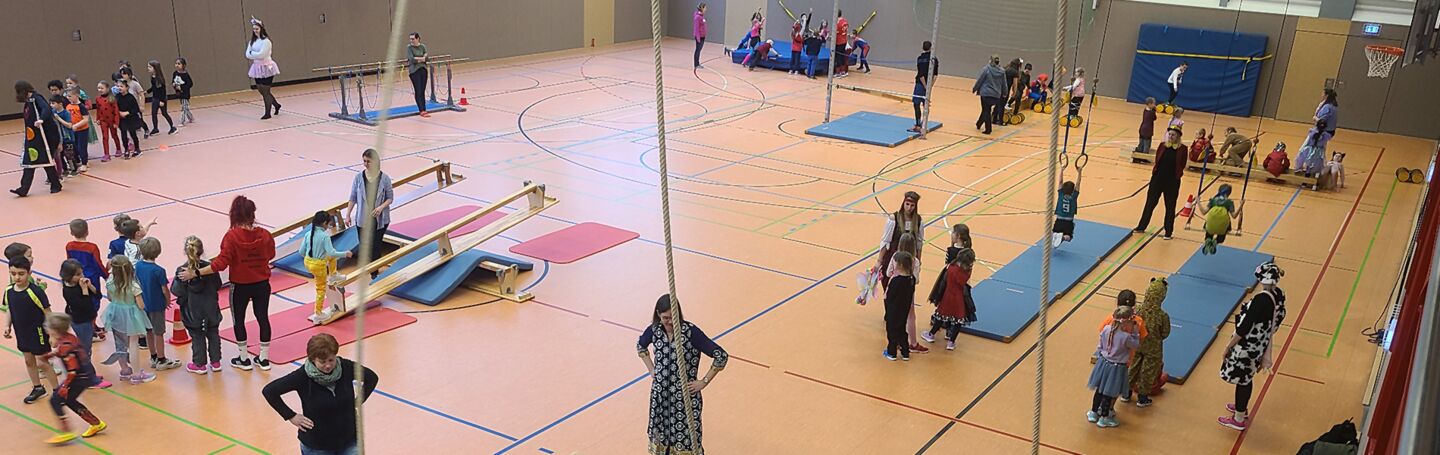 Eine Aufnahme vom Sportfest der Schule am Park. Man sieht eine große Turnhalle von innen mit verschiedenen Turngeräten und Matten. In der Turnhalle verteilt sind Schulkinder.